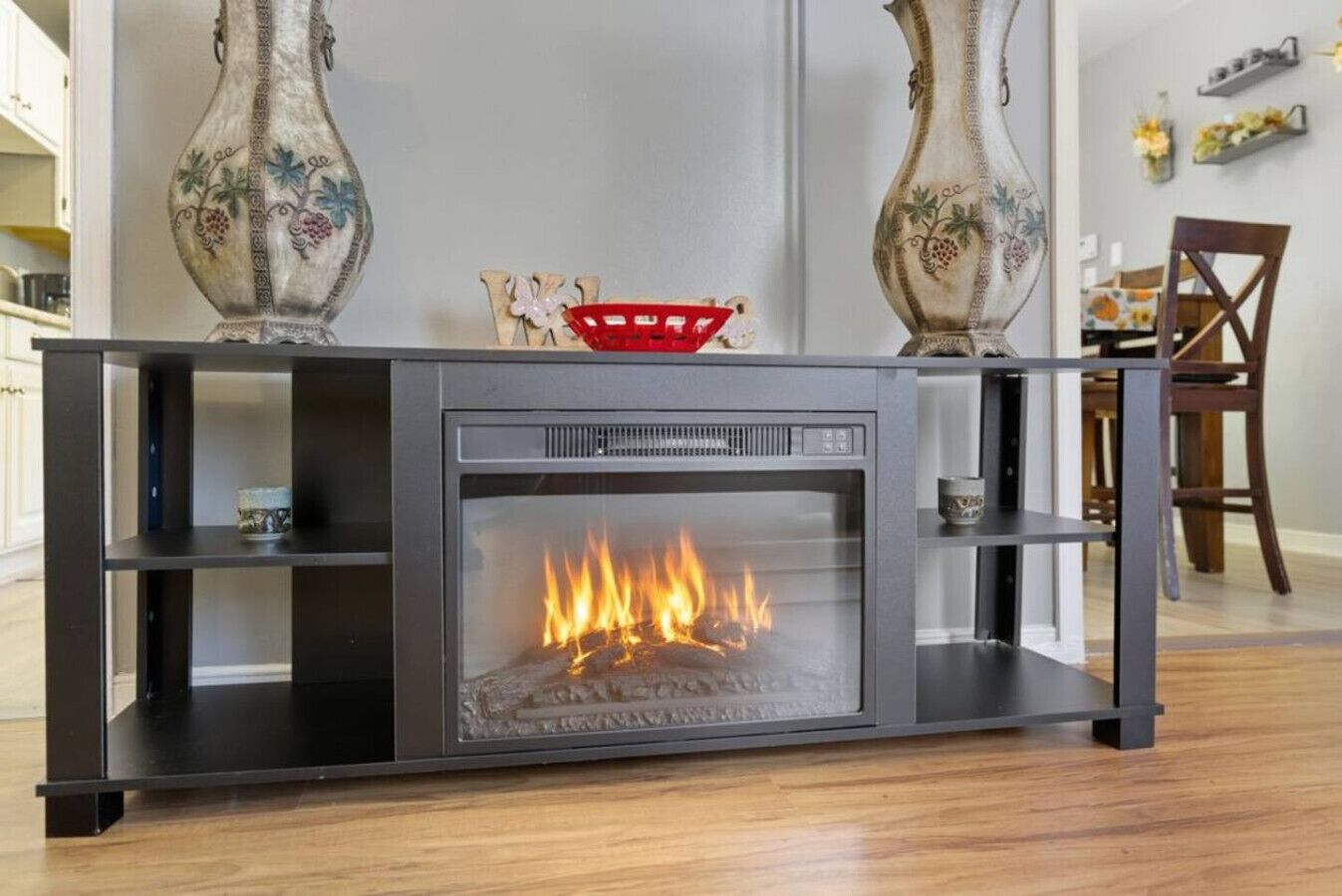 Una chimenea moderna con frente de vidrio, flanqueada por dos jarrones decorativos, en una habitación con piso de madera y un comedor al fondo.