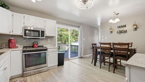 A modern kitchen with white cabinets, a stainless steel stove, and a red kettle. There is a dining area with wooden chairs and a table. - Next Stop: Comfort • 3 BR Near Amtrak Station (Richmond)