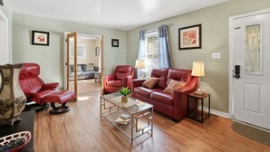 A living room with a red leather sofa, a glass coffee table, and a red armchair. There is a door with a decorative glass panel and a framed picture on the wall. - Next Stop: Comfort • 3 BR Near Amtrak Station (Richmond)