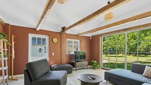 Ein Wohnzimmer mit einer Couch, einem Hocker und einem Couchtisch. Es gibt einen Fernseher, ein Fenster und eine Tür.