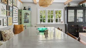 Ein Essbereich mit einem langen Tisch, Stühlen, einer hängenden Leuchte, einem Fenster mit Blick auf Grünflächen und einem grünen Schrank.