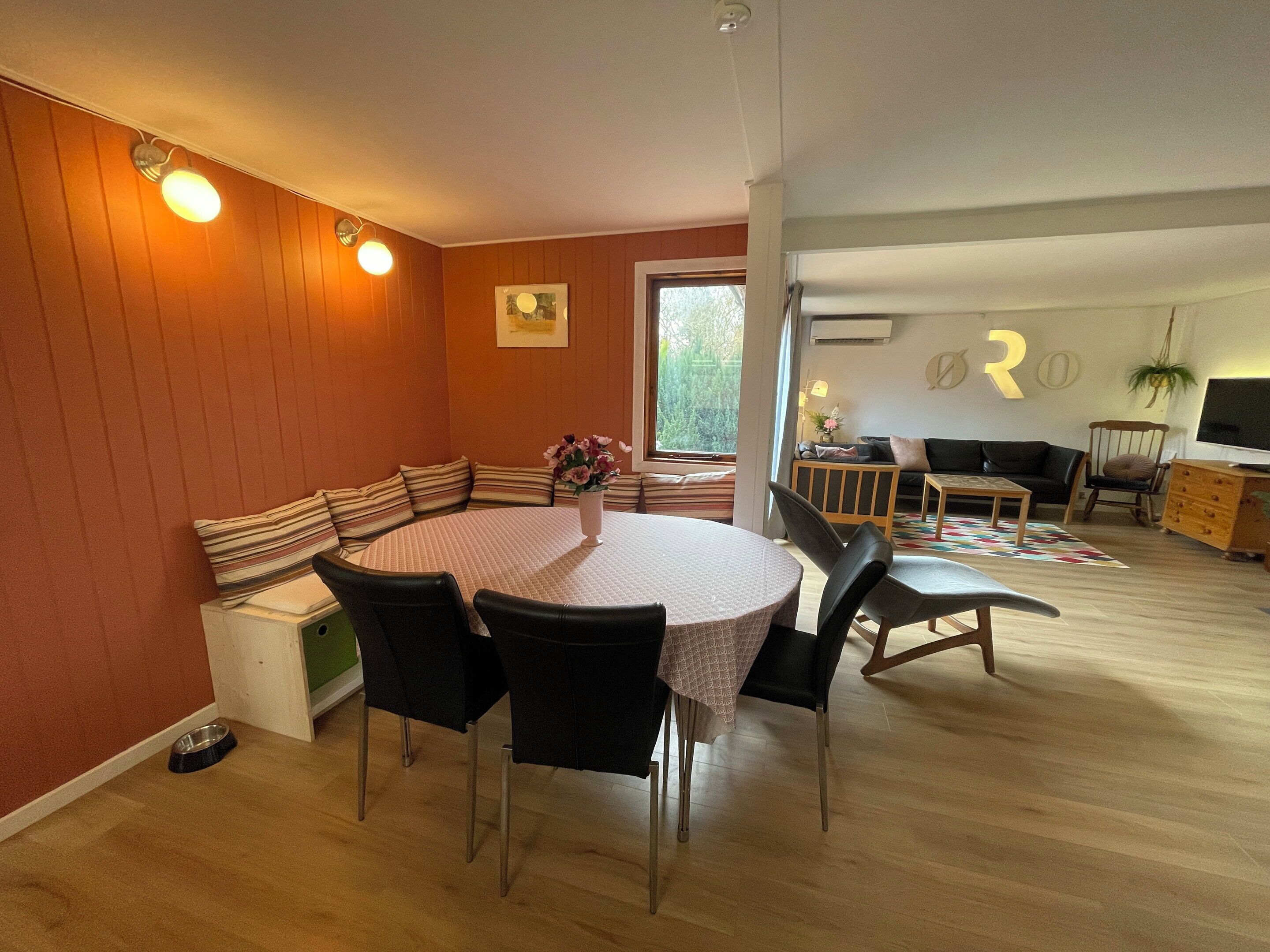 Een eetgedeelte met een ronde tafel, zwarte stoelen, een bank met gestreepte kussens, en een vaas met bloemen.