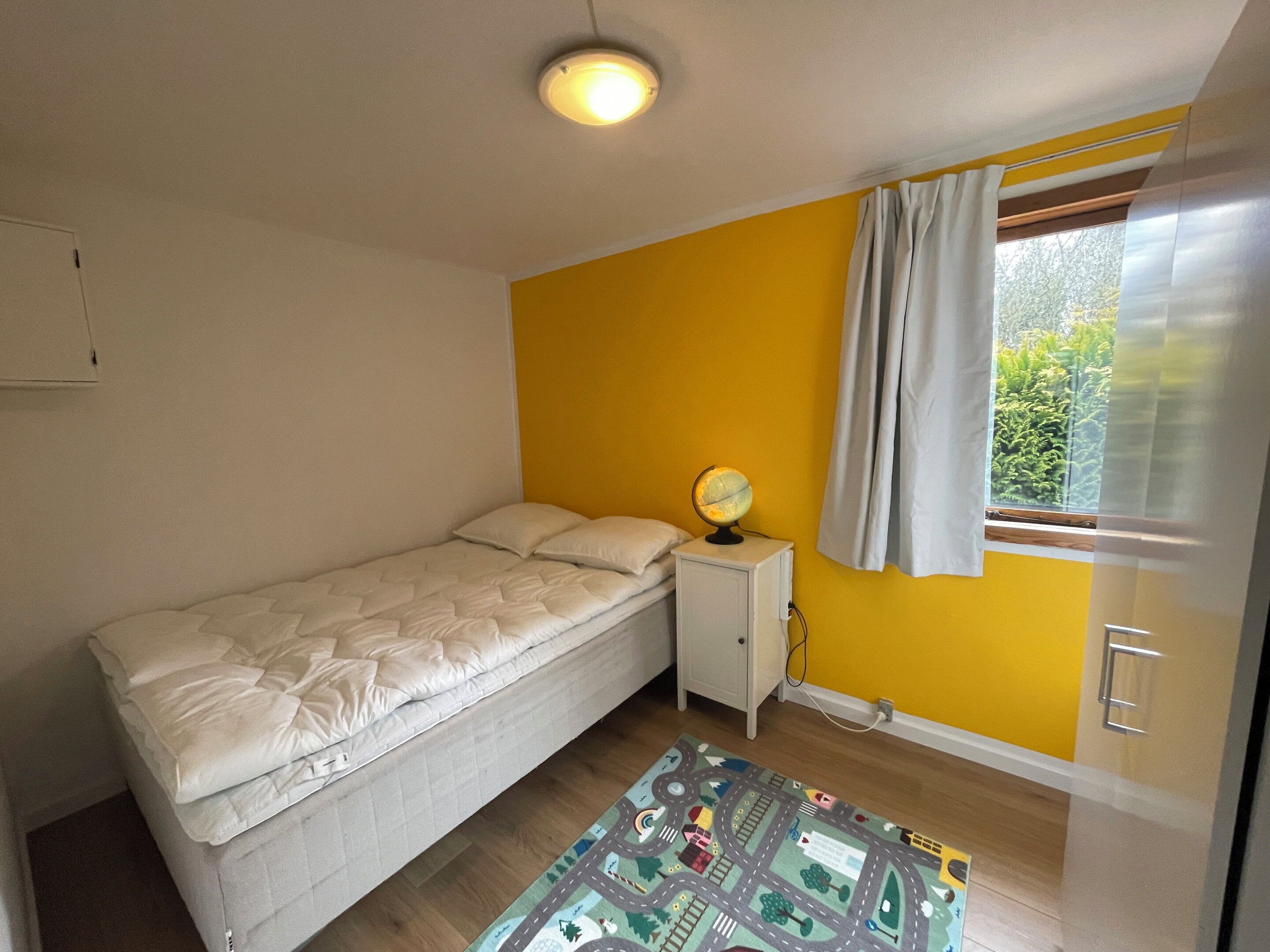 Une chambre à coucher avec un lit, une table de chevet sur laquelle se trouve un globe terrestre, une fenêtre avec des rideaux et une vue sur la verdure à l’extérieur.