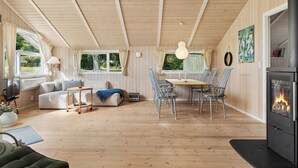 A cozy cabin interior with a wooden floor, a sofa, a dining table with chairs, a fireplace, and large windows. - Inviting summer house on scenic Røsnæs. (Kalundborg)