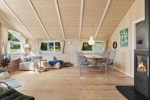Ein gemütliches Blockhaus-Interieur mit Holzboden, einer Couch, einem Esstisch mit Stühlen, einem Kamin und großen Fenstern.