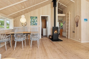 Un intérieur en bois comprenant un coin repas, un poêle en bois noir et une vue dégagée sur l’extérieur grâce à une porte ouverte.