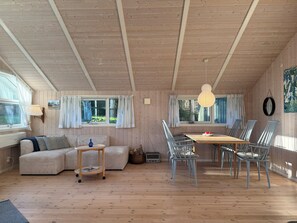 Een gezellig chaletinterieur met een houten plafond, een eettafel met stoelen, een bank en een kleine tafel met een lamp.