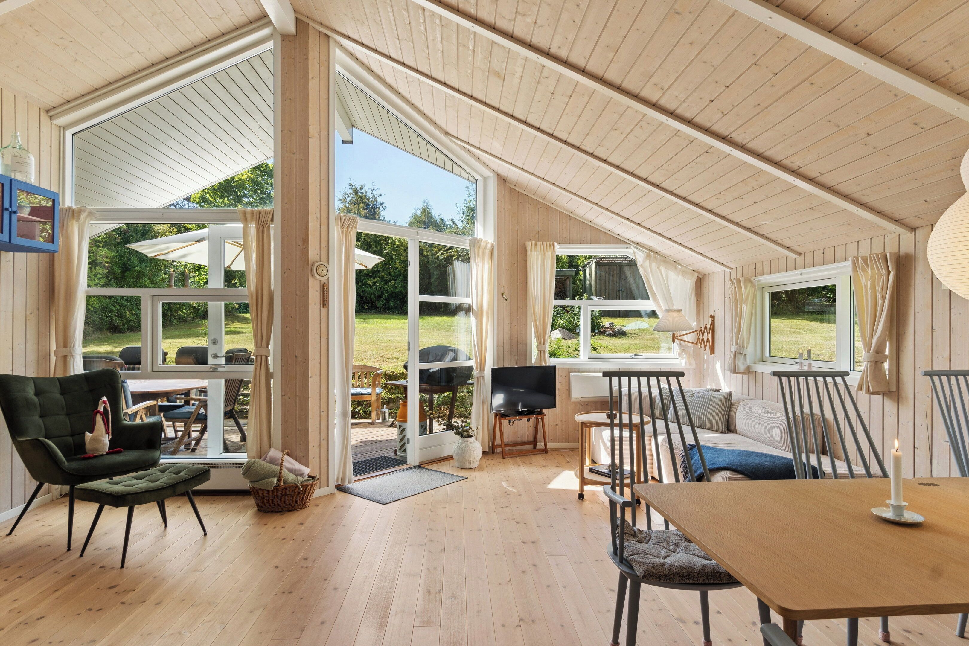 A spacious room with wooden flooring, a dining table, a sofa, and a TV. There are large windows and a door leading to an outdoor area.