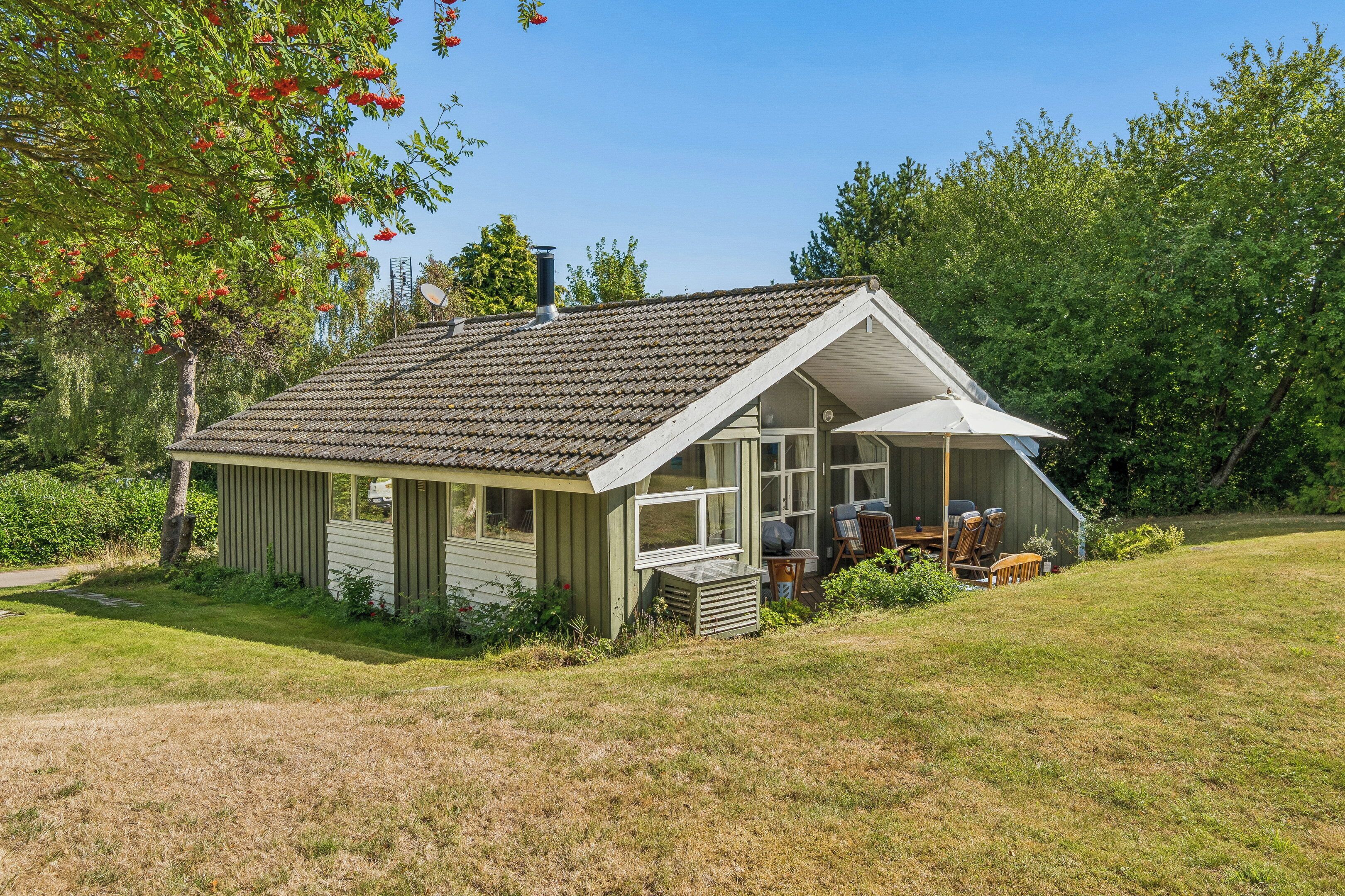A small, single-story house with a sloped roof, a porch with a table and chairs, and a garden area.