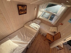 Eine kleine Holzhütte mit einem Einzelbett, einem Holzstuhl, einem Fenster mit Vorhängen und einem Gemälde an der Wand.