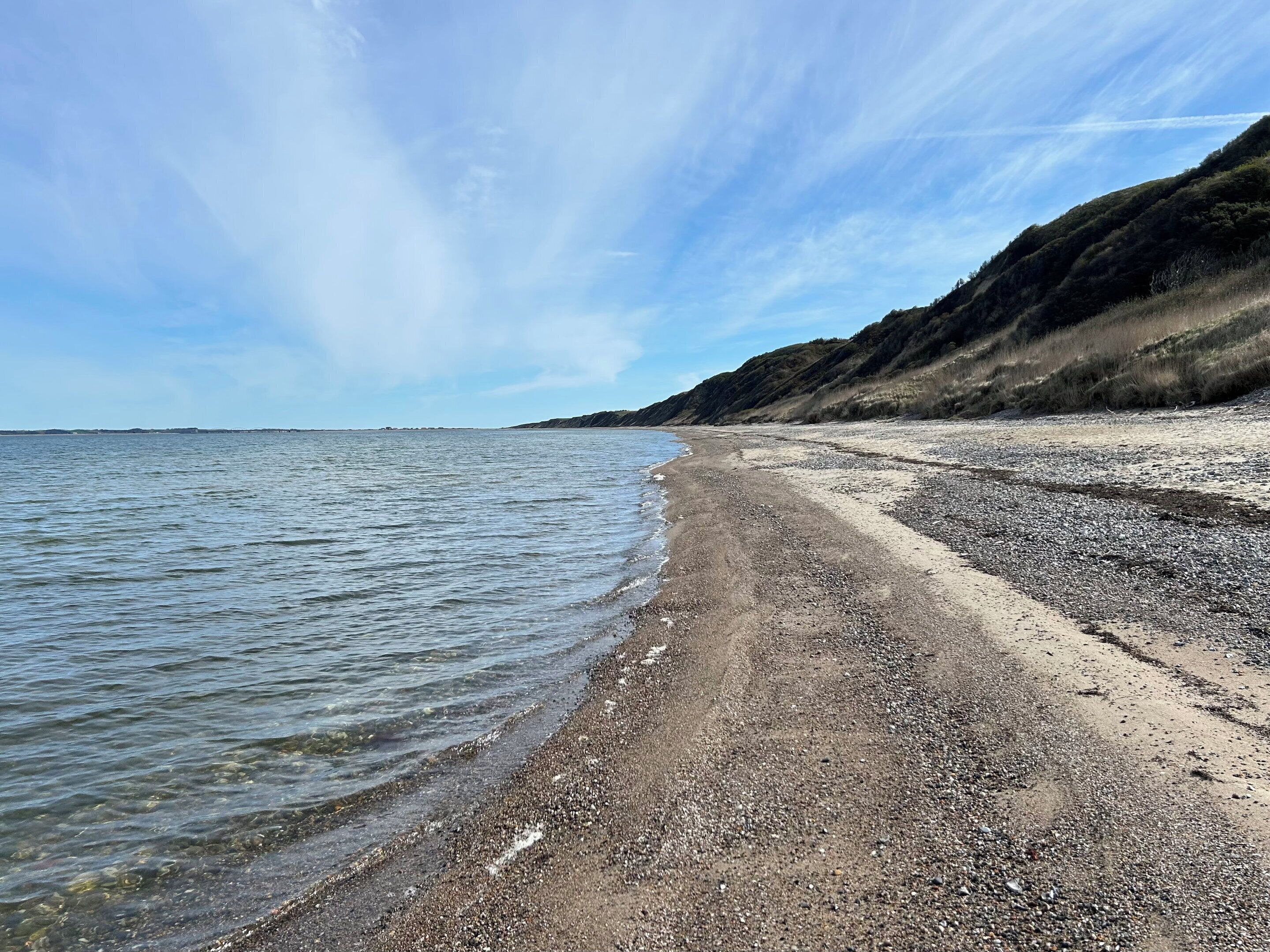 Ein Strand mit klarem Wasser, Kieselsteinen und einer hügeligen Landschaft im Hintergrund.