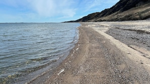 Une plage à l’eau claire, avec des galets et un paysage vallonné en arrière-plan.