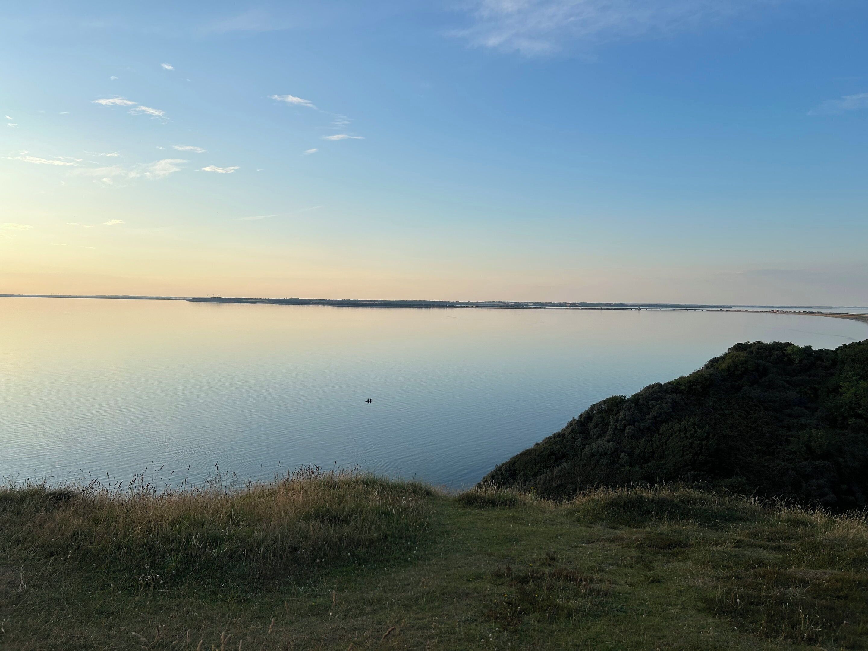Un plan d’eau paisible, avec un pont qui se dessine à l’horizon, un terrain herbeux et un ciel dégagé.
