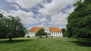 A large, white building with a red roof, surrounded by a well-maintained lawn and trees. - Hindemae Estate for holidays and events (Ullerslev)