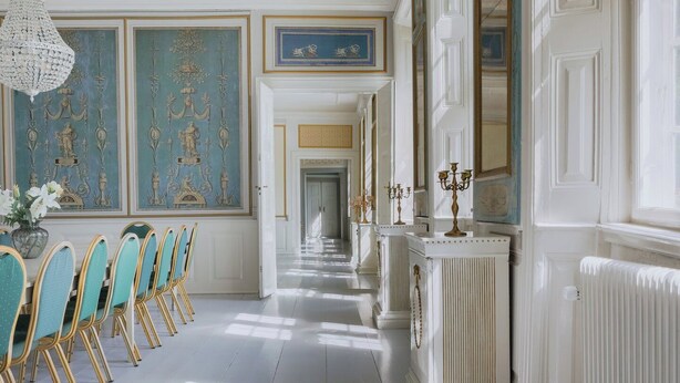 A long hallway with a series of chairs and a table, a radiator, and a mirror on the wall. - Hindemae Estate for holidays and events (Ullerslev)