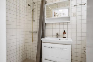 A bathroom with white tiled walls, a shower with a handheld showerhead, a sink with a bottle of hand soap, and a mirror above the sink. - Bright modern holiday home near Hindemae (Ullerslev)