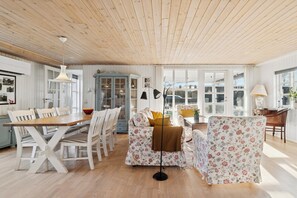 A room with a wooden ceiling, white walls, a dining table with chairs, a sofa, a floor lamp, and a cabinet. - Classic holiday home near beach and nature (Grenå)