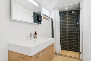 A modern bathroom with a white sink, wooden vanity, a mirror, and a shower area with dark tiles. - Classic holiday home near beach and nature (Grenå)
