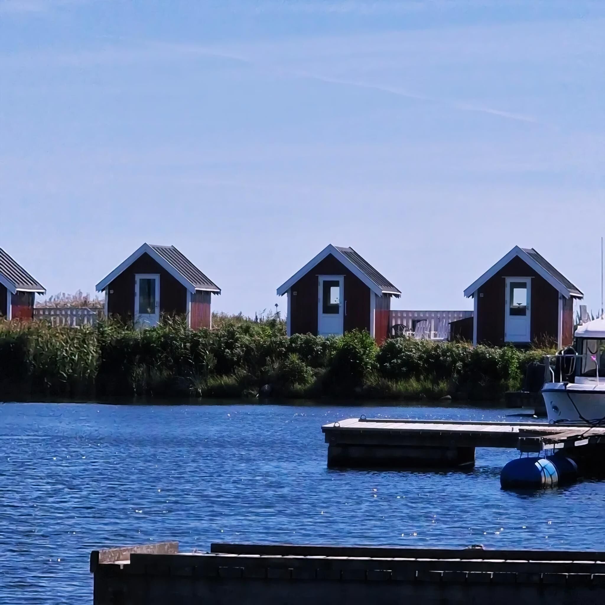 Three small, dark-colored cabins with pitched roofs, situated along a calm body of water, likely a lake or river, with a dock in the foreground.