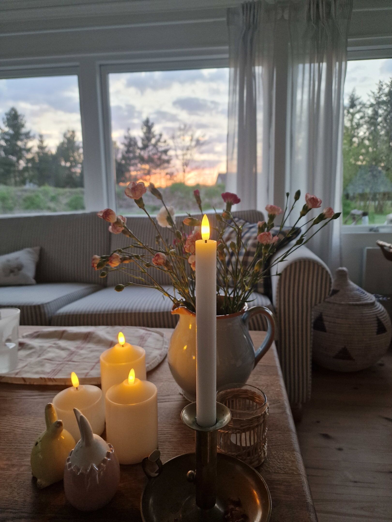 Een gezellige binnenshuis setting met een tafel waarop kaarsen staan, een vaas met bloemen en uitzicht door een raam.