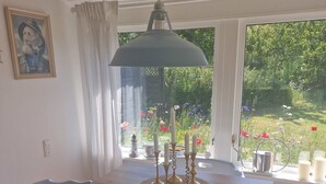 Een eetruimte met een houten tafel, stoelen, een hanglamp en uitzicht op een tuin door grote ramen.