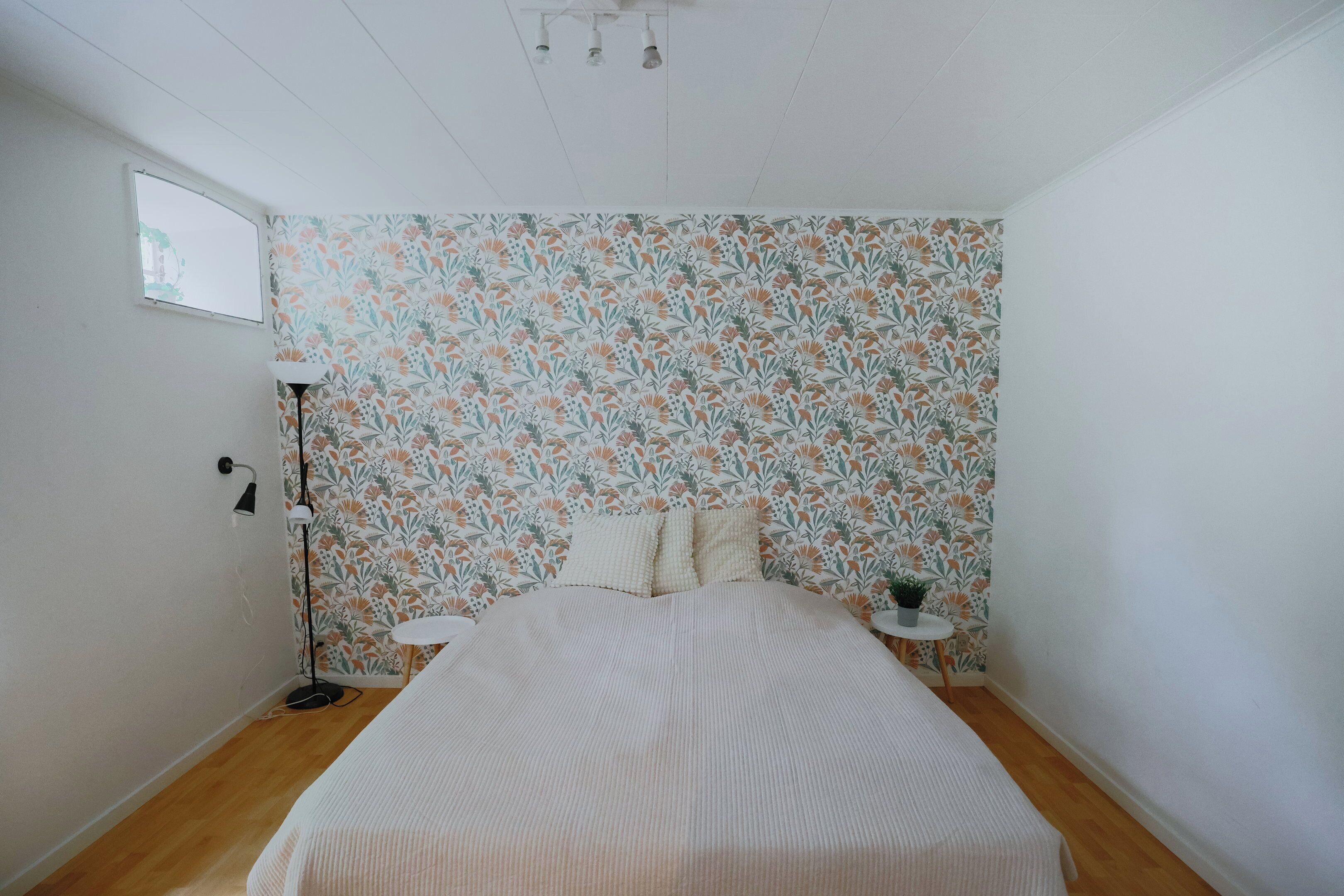 Une chambre avec un papier peint à motifs floraux, un lit recouvert d’une courtepointe à rayures, une table de chevet ornée d’une plante et une lampe sur pied.