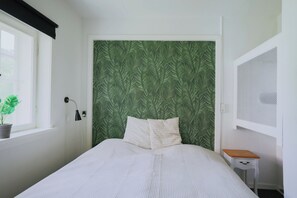 A bedroom with a bed, a window with a plant, a nightstand, and a wall with a leaf pattern. - Stay in Hindemae’s historic manager’s house (Ullerslev)