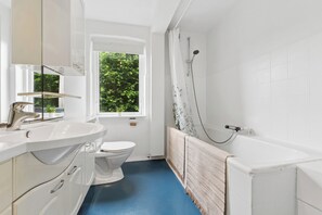 Un baño con lavamanos, inodoro y bañera blancos, una ventana con vistas a la vegetación y un suelo azul.
