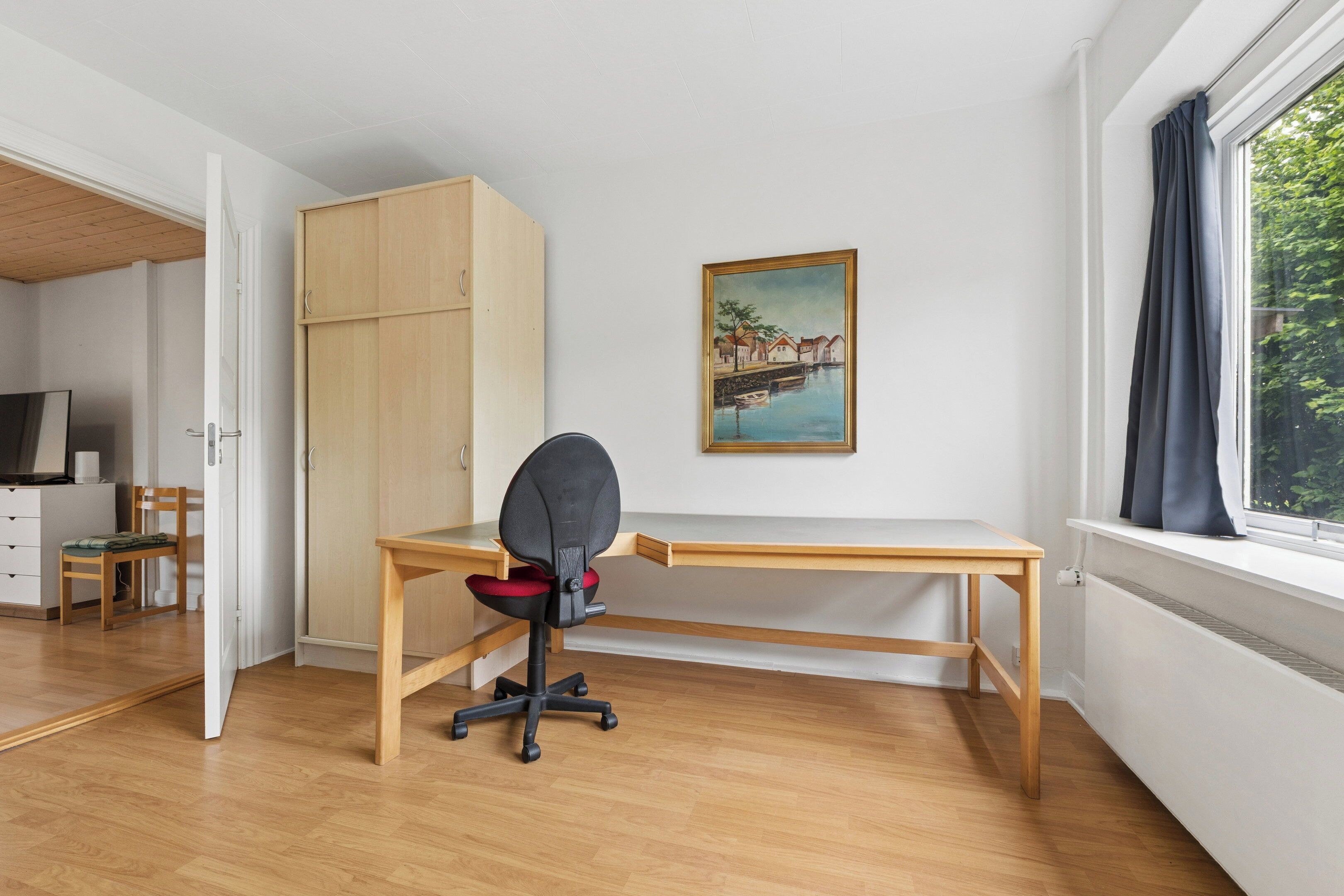 Et værelse med et træskrivebord, en stol, et garderobeskab, et billede og et vindue med gardiner.