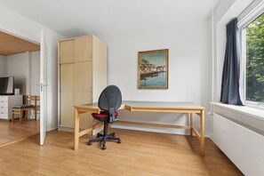 A room with a wooden desk, a chair, a wardrobe, a painting, and a window with curtains.