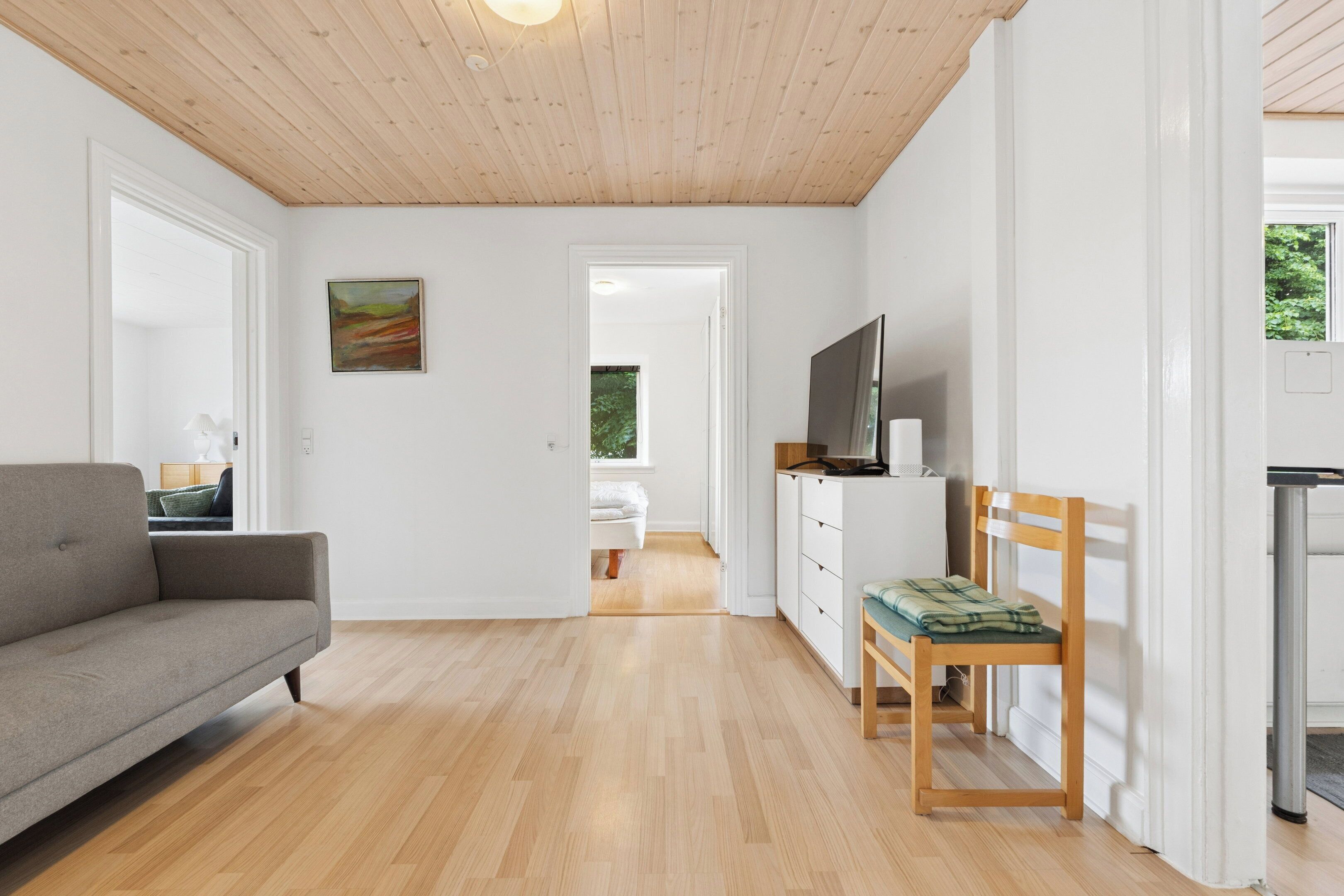 Een moderne woonkamer met een bank, een televisie, een houten stoel en een uitzicht op een slaapkamer door een geopende deur.