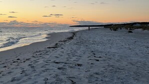 En strand med fotspår som leder ner mot vattnet, en person som går och en solnedgång i bakgrunden.