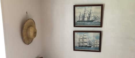 Ein Zimmer mit einem Holzstuhl, einem an der Wand befestigten Hut und zwei gerahmten Bildern von Segelschiffen.