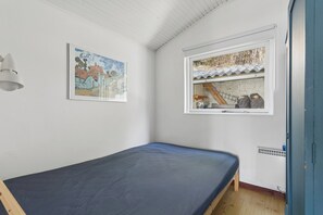 Ein Einzelbett mit blauer Matratze in einem Zimmer mit weißen Wänden, einem Fenster mit Blick auf ein Haus und einem gerahmten Gemälde an der Wand.