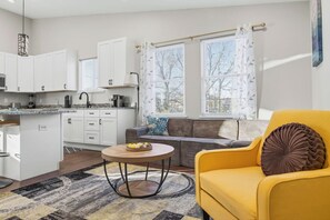 Un espacio habitable compacto con kitchenette, un sofá, un sillón amarillo y una mesa redonda.