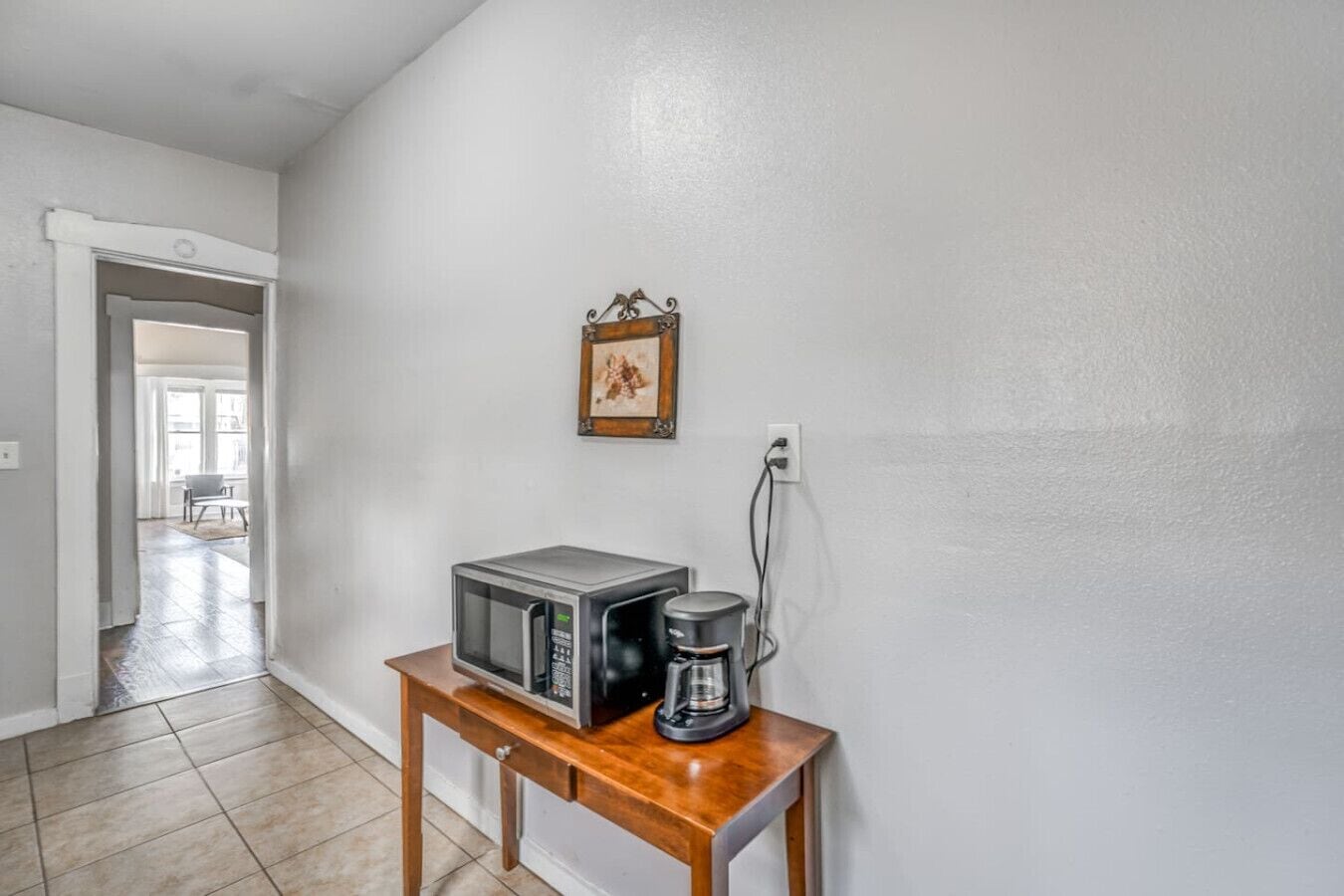 Una zona de cocina con microondas y cafetera sobre una mesa de madera, un enchufe y un cuadro en la pared.