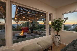 Un salon chaleureux avec une grande fenêtre donnant sur un patio agrémenté d’un foyer et de guirlandes lumineuses.