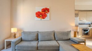 A modern living room with a grey sofa, a painting of red flowers, and a kitchen in the background. - Kalives Sand - Beachfront apartment (Chania)