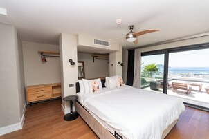 Un dormitorio moderno con una cama grande, un ventilador de techo, una mesita de noche pequeña, un armario y un balcón con vistas al mar.