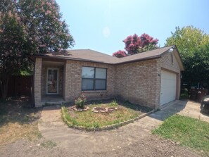 A single-story brick house with a front garage, a small front yard with a flower bed, and a tree with pink flowers. - San Riveria Rentals-Cozy Pool property ! Enjoyable 3-bedroom house with AC in SA (San Antonio)