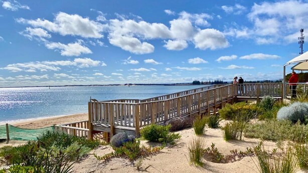 Un paseo marítimo de madera que lleva a una playa, con vista clara al agua y la costa distante.