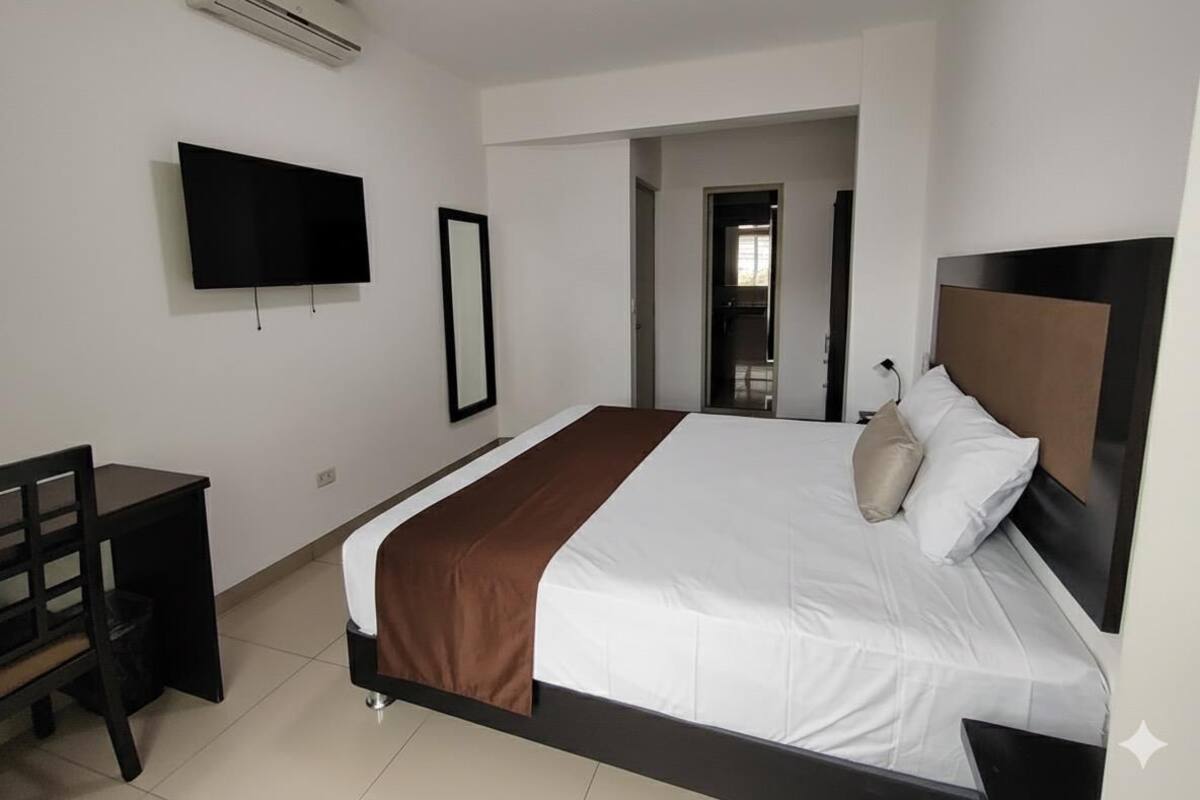 A modern hotel room with a large bed, a desk, a flat-screen TV, and a door leading to a bathroom.