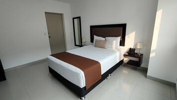 Quarto de hotel com uma cama grande, uma mesa de cabeceira com abajur e uma porta.