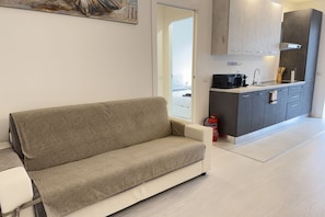 En moderne stue med sofa, et køkken med lysegrå skabe og en synlig brandslukker.