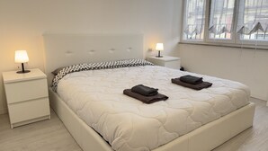 Una cama bien hecha con colcha, dos almohadas y toallas dobladas sobre un cabecero blanco. Hay dos mesitas de noche con lámparas y una ventana con cortinas.