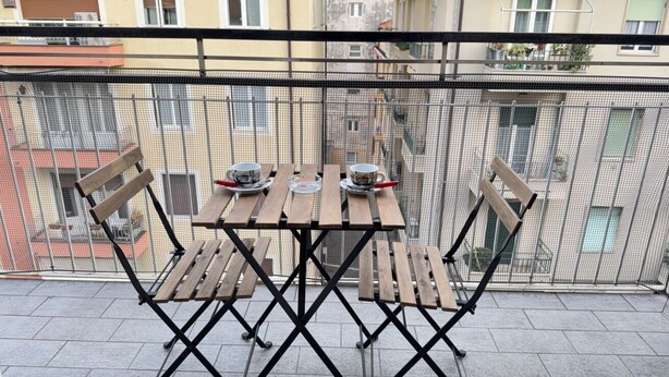 Un balcón con mesa y sillas, una barandilla de metal y edificios al fondo.