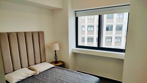Ein Schlafzimmer mit einem Bett, einem Nachttisch, einer Lampe und einem Fenster mit Blick auf Gebäude.