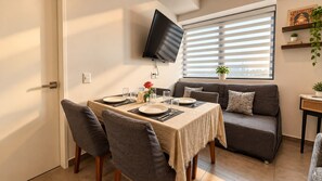 Ein Essbereich mit einem Tisch für zwei Personen, einer Couch, einem an der Wand befestigten Fernseher und einem Fenster mit Jalousien.