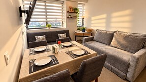Ein Essbereich mit Tisch für vier Personen, einem grauen Sofa und einem Fenster mit Jalousien.
