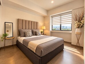 A modern bedroom with a large bed, a bedside table with a lamp, a window with blinds, and a potted plant. - App. Lafayette 10A: 2 bedrooms, 2 bathrooms, 1 parking space (Guadalajara)